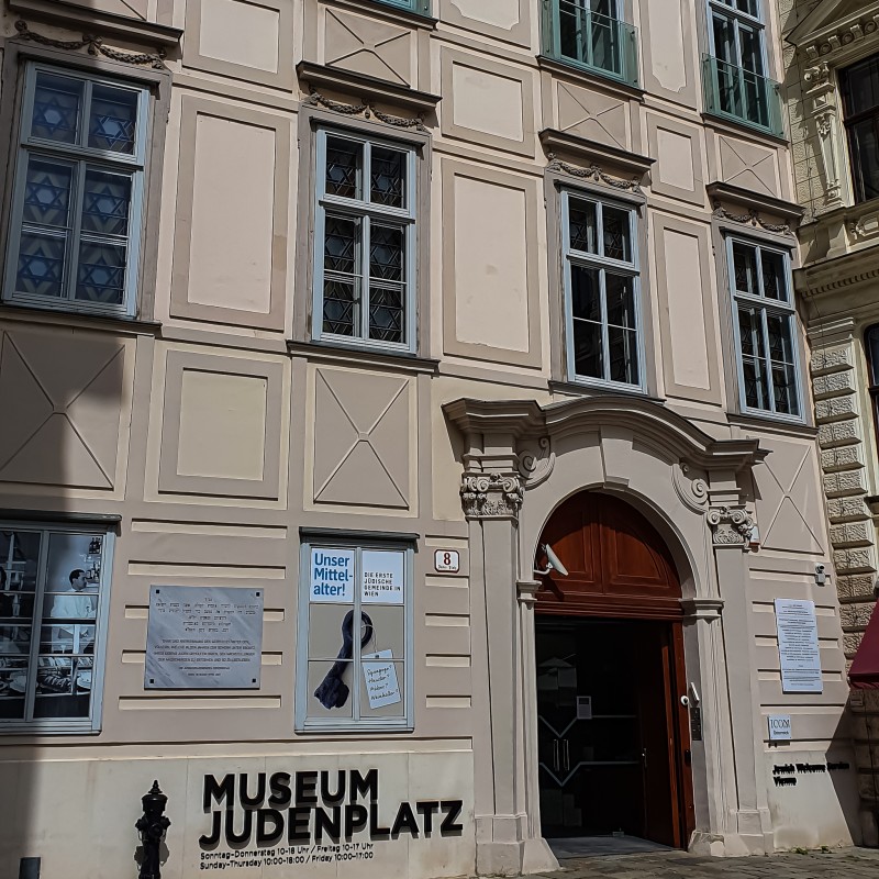Museum Judenplatz facade: Facade of the Museum Judenplatz with five windows and a dark entrance under an ornately decorated arch, with signs and posters on the building