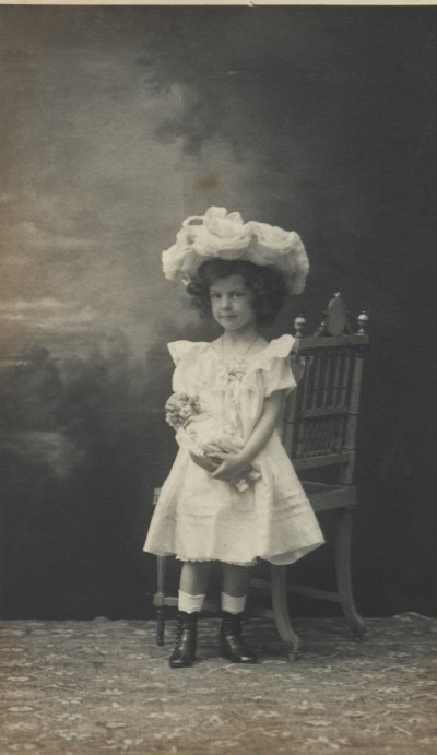 Portrait of a girl with doll: Girl in white dress and large floral hat stands beside a chair holding a doll.