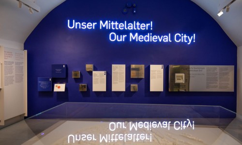 Ausstellungstafel mit dem leuchtenden Schriftzug ‚Unser Mittelalter! Our Medieval City!‘ und Informationstafeln an einer dunkelblauen Wand