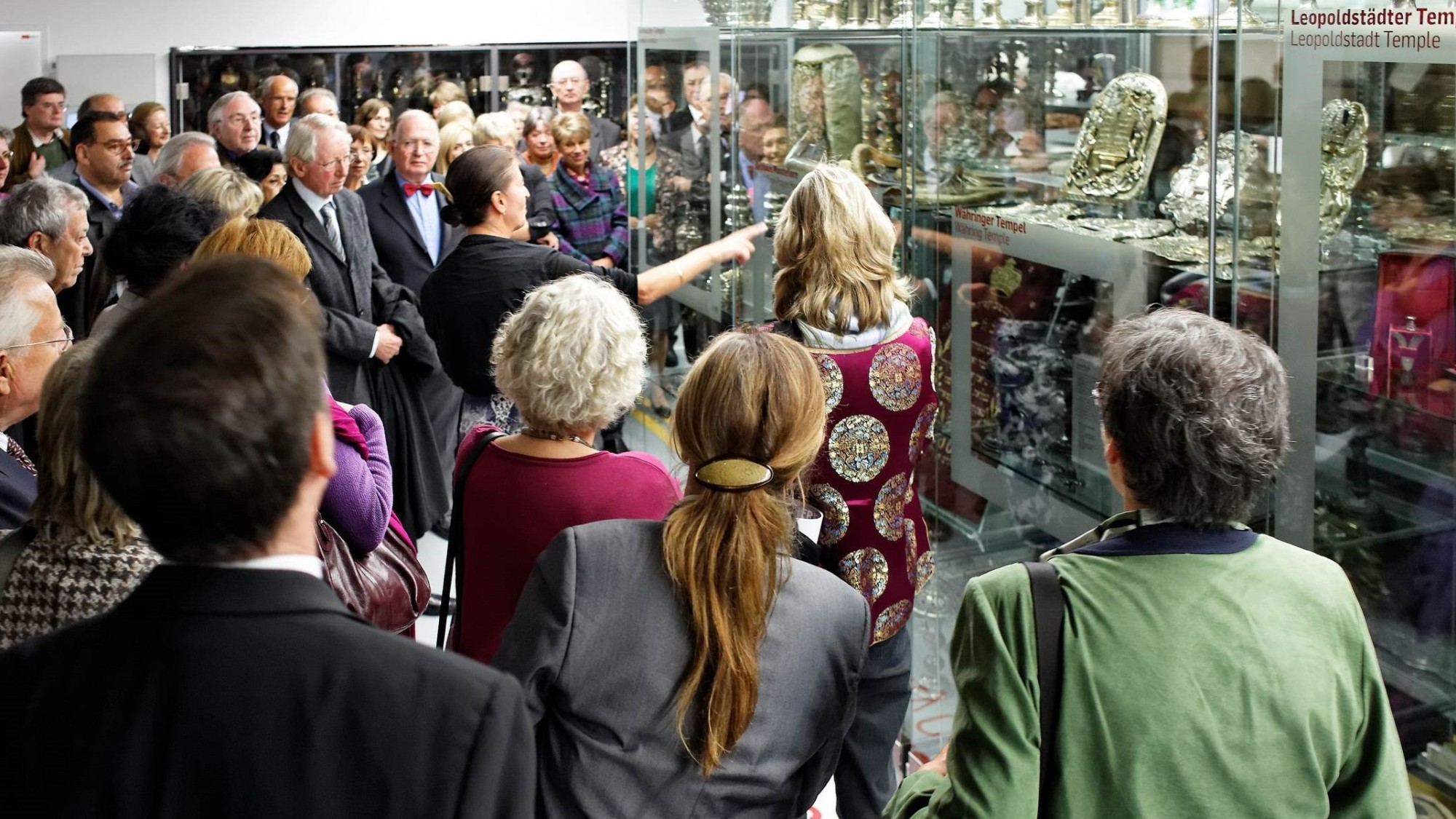 Tour at the Leopoldstadt Temple Museum: Group of adults attending a museum tour in front of glass cases displaying silver and metal artifacts, sign reading 'Leopoldstadt Temple' visible