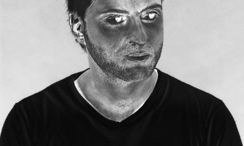 Junger Mann mit kurzem dunklem Haar, Bart und ernstem Blick, trägt ein schwarzes T-Shirt mit V-Ausschnitt