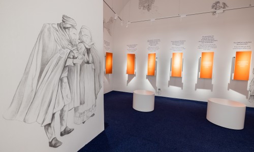 Wandzeichnung von zwei historischen Figuren in Umhängen links, rechts sechs orange beleuchtete Informationstafeln mit Text in Deutsch und Englisch, zwei weiße Sitzhocker auf blauem Boden.