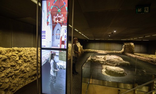 Digitale Anzeige mit virtueller Darstellung einer historischen Kirche neben antiken Mauerresten in einem Museumssaal