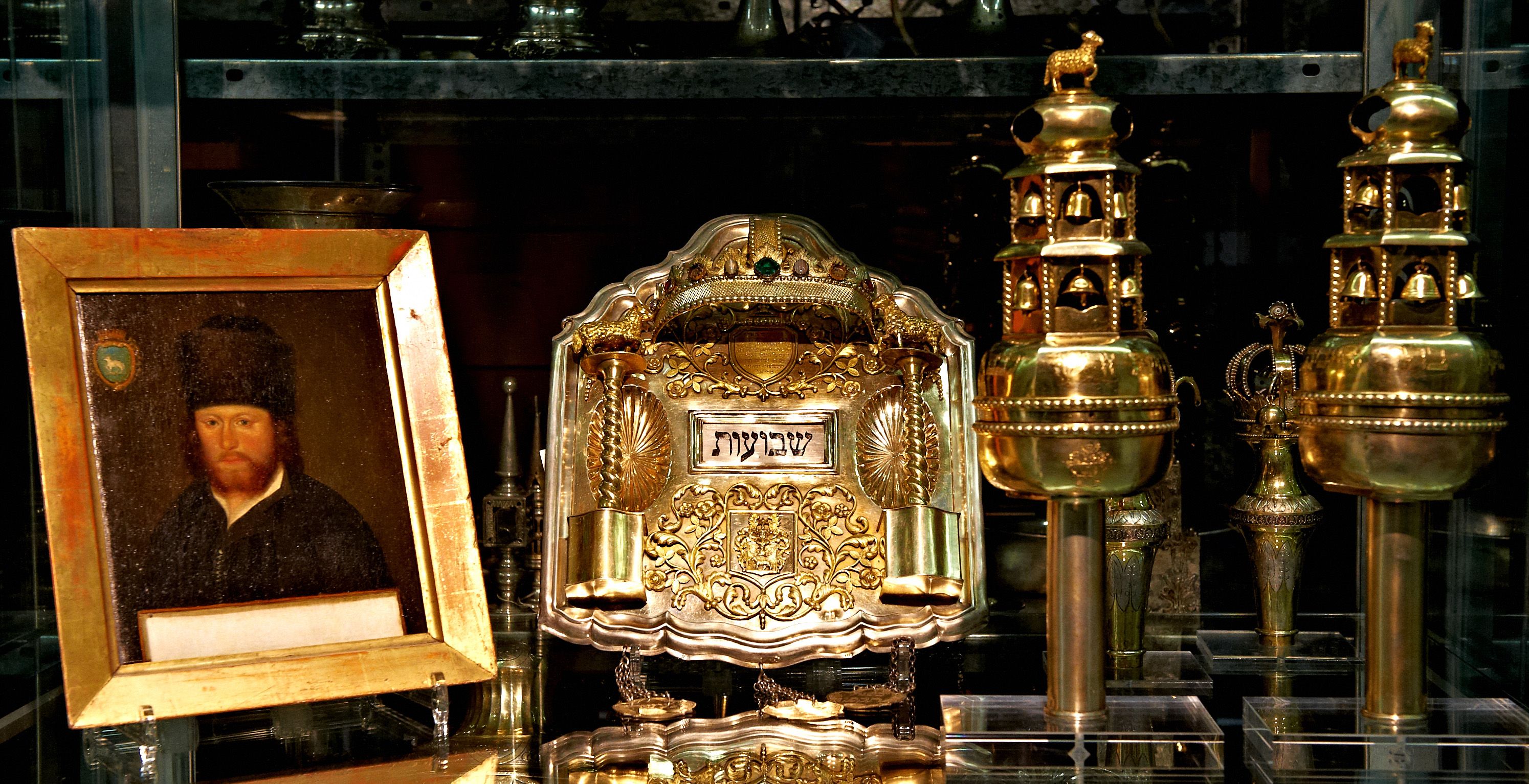Porträt eines Mannes mit Bart und Pelzmütze neben einem silbernen Schriftrollenhalter mit hebräischer Inschrift und zwei goldenen Toraschmuckstücken mit Glocken in einer Vitrine