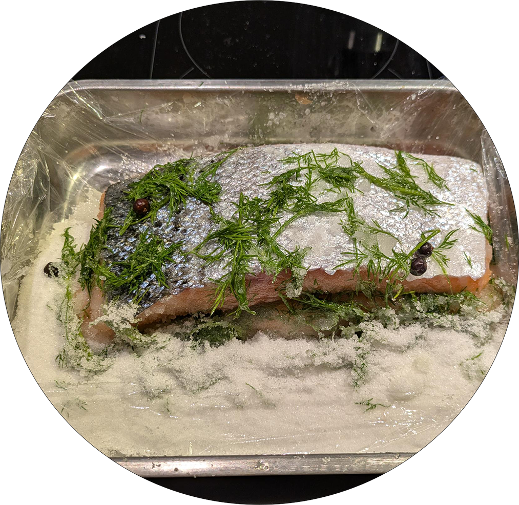 Salmon piece with skin in a metal tray on a bed of salt and dill with juniper berries.
